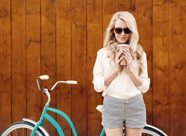 Joven chica rubia sexy con el pelo largo en gafas de sol de pie cerca de la bicicleta verde vintage y la celebración de una taza de café divertirse y buen humor mirando en la cámara y sonriendo, cálido, tonificación Imagen de stock