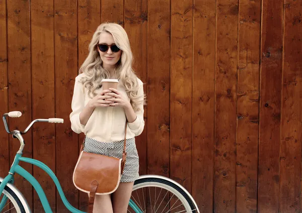 Joven chica rubia sexy con el pelo largo con bolso vintage marrón en gafas de sol de pie cerca de la bicicleta verde vintage y sosteniendo una taza de café, divertirse y buen humor mirando en la cámara y sonriente, cálido , Imágenes de stock libres de derechos