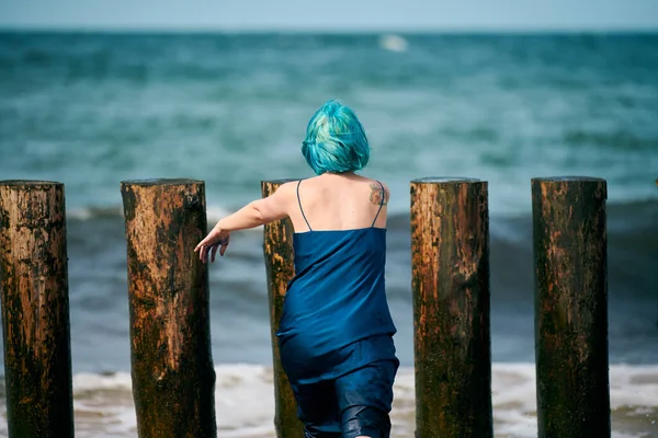 Beautiful blue-haired woman performance artist in dark blue dress standing on beach holding paint brush, back view. Contemporary abstract art, art of dancing in nature, modern performance art outdoor