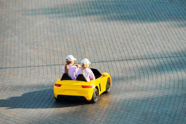İki küçük kız lunaparkta kaldırımda sarı oyuncak araba kullanıyor. Çocuklar uzaktan kumandalı elektrikli araba kullanıyorlar. Çocuk eğlencesi için pil aracı