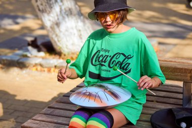 Zelenogradsk, Rusya - 07.30. 2021 - Çelik dilli davul çalan sokak müzisyeni kız. Hank davul dinlendirici sokak müziği. Panama giymiş hoş bir kız ve yeşil tişörtlü. Şehir caddesinde davul çalıyor.