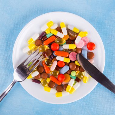 Beyaz bir tabakta çatal ve bıçakla çeşitli renkli ilaçlar ve haplar.