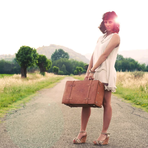 Hermosa chica esperando en un camino de campo con su maleta — Foto de Stock