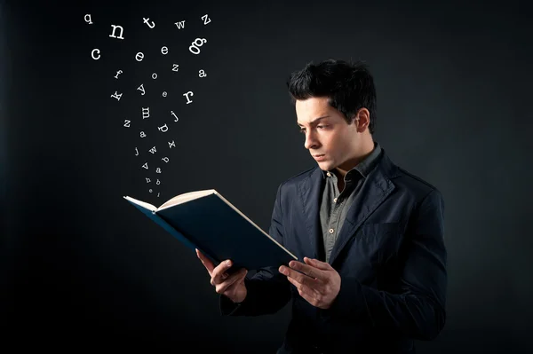 Young man reading book with letters coming out against dark background — Stock Photo, Image