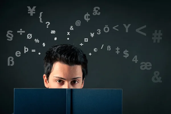 Young man behind a book with letters coming out of his head against dark background — Stock Photo, Image