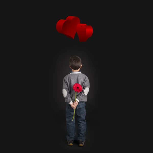 Child holds red flower behind his back. Black background with red hearts — Stock Photo, Image