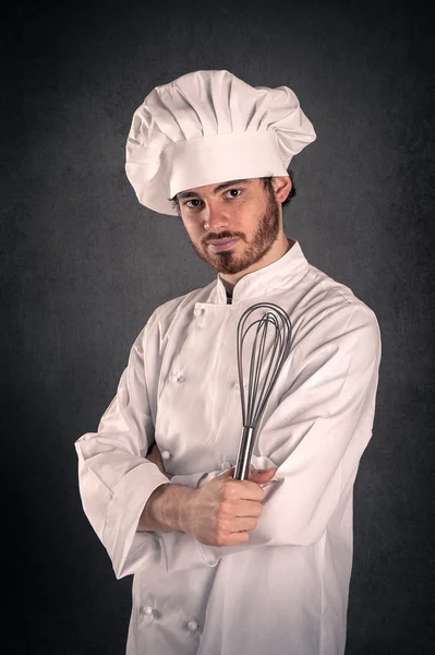 Portrait of a young cook man wearing uniform holding a steel whisk over grunge background. — Stock Photo, Image