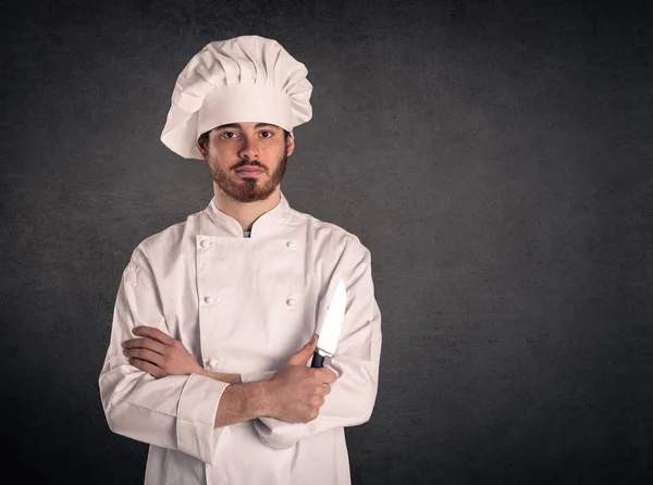 Retrato de un joven cocinero con uniforme sosteniendo un cuchillo sobre fondo grunge . — Foto de Stock