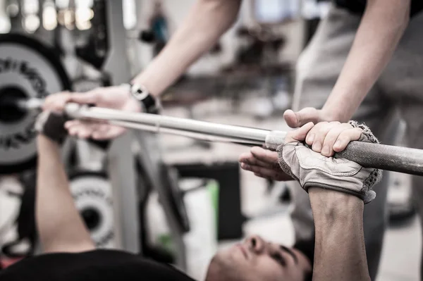 Joven levantando la barra en el gimnasio con instructor. Céntrate en la mano . — Foto de Stock