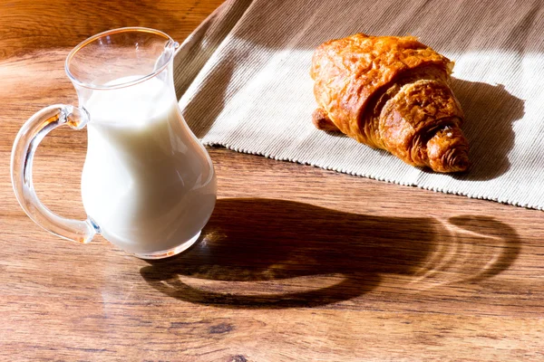 Glass jug of milk with croissant on wooden table — Stock Photo, Image