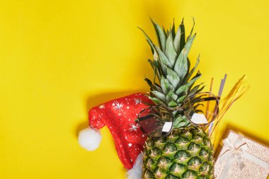 ananas, noel baba şapkası ve güneş gözlükleri, sarı arka planda noel dekoru, tropik kuşakta kış tatili