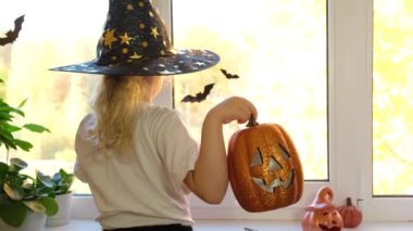 Cadı şapkalı, sarı saçlı şirin beyaz kız eğleniyor ve cadılar bayramını kutlamak için pencereyi süslüyor. Penceredeki cadılar bayramı dekorasyonu