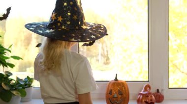 Cadı şapkalı, sarı saçlı şirin beyaz kız eğleniyor ve cadılar bayramını kutlamak için pencereyi süslüyor. Penceredeki cadılar bayramı dekorasyonu