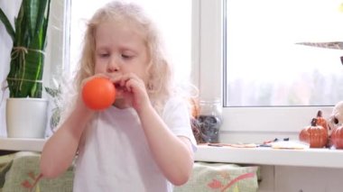 Sarı kıvırcık saçlı sevimli bir kız cadılar bayramı için bir balon şişiriyor, arka planda bitkilerle dolu bir oda ve cadılar bayramı dekoru var.