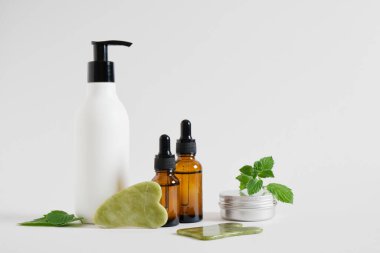 aluminum cosmetics jar, cream in a bottles with a dispenser, serum in dropper bottle and a gua sha scraper made of jadeite stone, mint leaves on gray background clipart