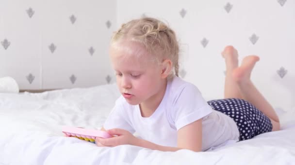 Uma menina de 5-6 anos está deitada em uma cama e joga smartphone, pop it smartphone case, criança e conceito de gadgets — Vídeo de Stock