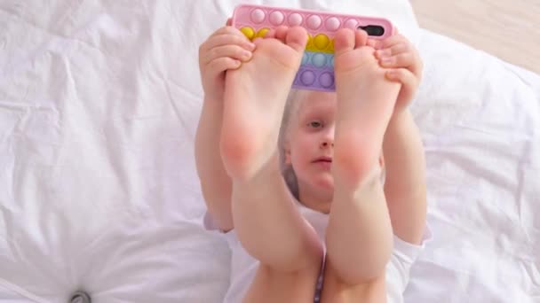 Une fille de 5-6 ans est allongée sur un lit et joue au smartphone, pop it cas de smartphone, enfant et gadgets concept — Video