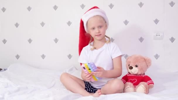 산타 클라우스 모자를 쓴 소녀가 침대에 앉아 있는 동안 스마트폰을 가지고 노는 모습 — 비디오