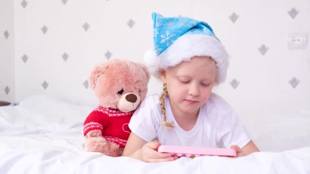 산타 클라우스 모자를 쓴 한 소녀가 침대에 누워 스크린 스마트 폰을 보고 있습니다. — 비디오