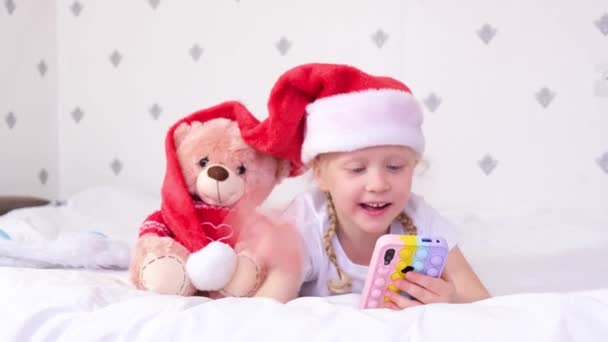 Una chica en un sombrero rojo de Santa Claus mira a la pantalla de un teléfono inteligente y se encuentra en la cama con una mano agitada — Vídeo de stock