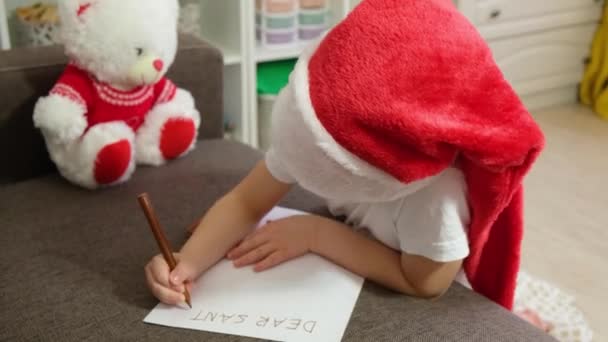 Schattig Kaukasisch meisje 5-6 jaar oud in een santa hoed schrijft een brief aan de kerstman — Stockvideo