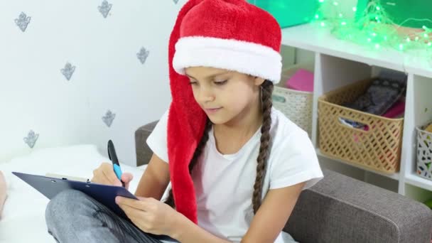 可爱的棕色头发的小女孩戴着红色的桑塔帽，给坐在床上的桑塔 · 克洛斯写信 — 图库视频影像