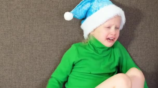 Llorando linda niña de 5-6 años en un suéter verde y un sombrero de Santa Claus sentado en el sofá, — Vídeo de stock