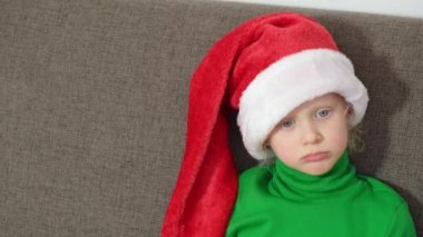 Üzgün, tatlı kız 5-6 yaşlarında, yeşil kazaklı ve Noel Baba şapkalı, kanepede oturan, üzgün ve kırmızı şapkalı.