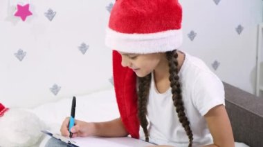 Kırmızı Noel Baba şapkalı tatlı kahverengi saçlı kız Noel Baba 'ya bir mektup yazıyor.