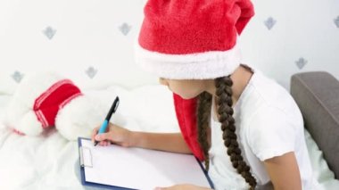 Kırmızı Noel Baba şapkalı tatlı kahverengi saçlı kız Noel Baba 'ya bir mektup yazıyor.