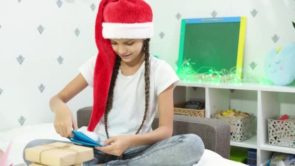 可愛いです茶色の髪の女の子ティーンでA赤サンタ帽子ベッドの上に座っているサンタクラスへの手紙を書きます クリスマスの雰囲気と休日のための準備 — ストック動画
