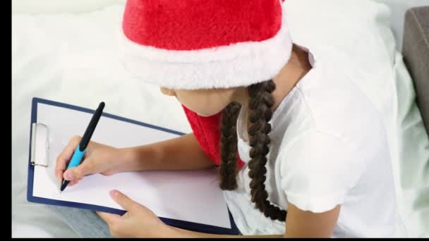 可愛いです茶色の髪の女の子ティーンでA赤サンタ帽子書きます手紙へサンタクラス座っているベッドの上に — ストック動画