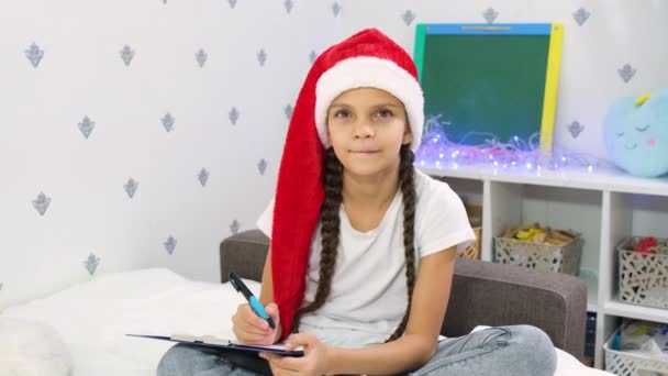 可愛いです茶色の髪の女の子ティーネージャーでA赤サンタの帽子は ベッドのクリスマスの装飾に座っているサンタクラスに手紙を書きます — ストック動画