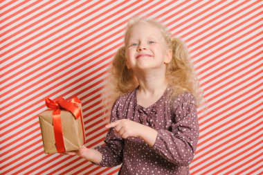 Kırmızı çizgili bir geçmişi olan neşeli anaokulu kızı, sarı saçlı bir kızın elinde kırmızı kurdeleli bir hediye, boks günü, doğum günü ve noel hediyeleri, tatil hediyeleri.