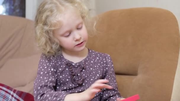 Roztomilý dívka s kudrnaté blond vlasy hrát s pop it trubka hračka sedí na židli — Stock video