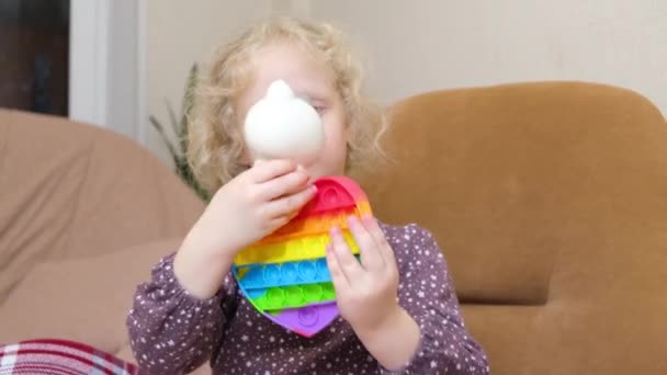 Niedliches Mädchen mit lockigem blondem Haar, das mit einem Pfeifenspielzeug spielt, das auf einem Stuhl sitzt — Stockvideo