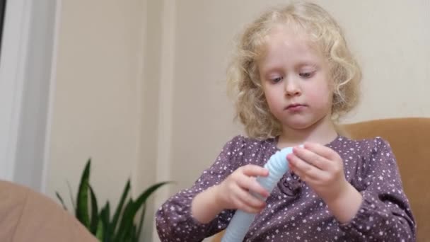 Linda chica con pelo rubio rizado jugando con pop it juguete de tubería sentado en una silla — Vídeos de Stock