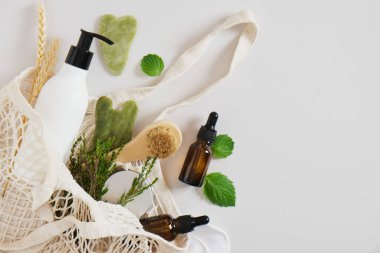 Vücut ve cilt bakımı için gri arka planda bir sicim torbasında, gua sha kazıyıcıları ve cilt bakımı, krem, losyon ve masaj yağı için kozmetik ürünleri, aromaterapi için nane yaprakları.