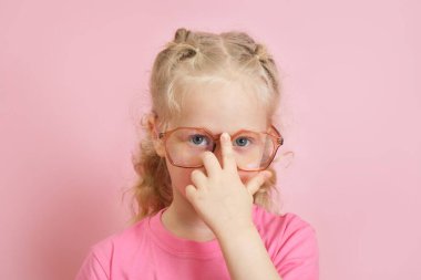 Pembe tişörtlü, burnuna gözlük takan beyaz bir anaokulu öğrencisinin portresi, çocuk göz doktoru konsepti, okula geri dönüyor.