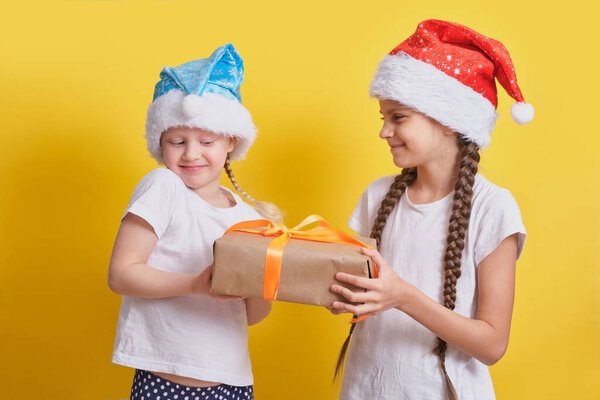 Happy smiling girls in festive Santa hats with christmas gift box on yellow background