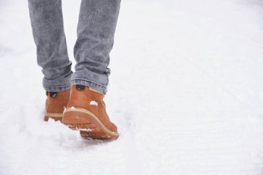 Kot pantolonlu ve deri çizmeli bir adamın bacakları karlı yolda yürür.