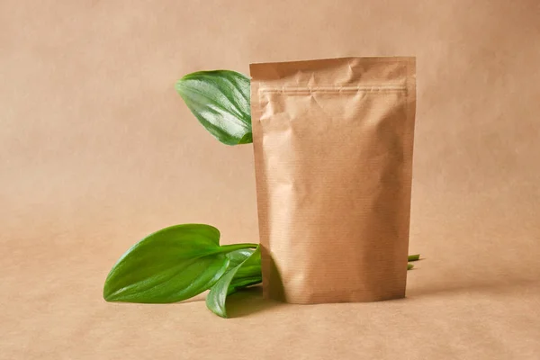 blank mockup paper bag packaging and green fresh leaves on brown background Eco friendly packaging, paper recycling, zero waste, natural products concept. Copy space.