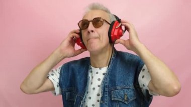 elderly gray-haired man in denim vest listens to music in retro collars on pink background, man enjoys music, feels young