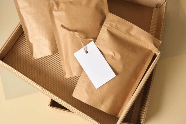 mock up blank paper bags with white tag for marking in corrugated cardboard box on beige background Eco friendly packaging, paper recycling, zero waste, natural products concept. Copy space.