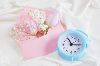 Hassas gölgelerde bir buket bahar çiçeği, bir çalar saat ve yatağın üzerinde pembe bir zarf, bir aşık için romantik bir hediye, 8 Mart ya da 14 Şubat sabahı ruh hali