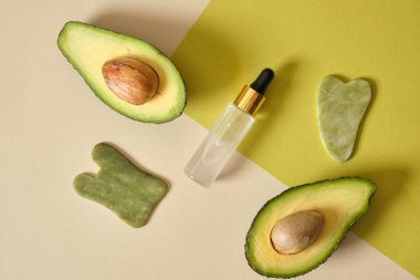 Jadeite gua sha kazıyıcılar ve taze ve dilimlenmiş avodcado meyveleri bej ve yeşil arka plan görünümlü avokado yağı kavramlı üst görünüm masajı