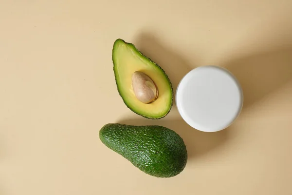 white mock-up cosmetics jar and avocado on a beige background, skin care with natural cosmetics, masks and creams with avocado oil copy space top view