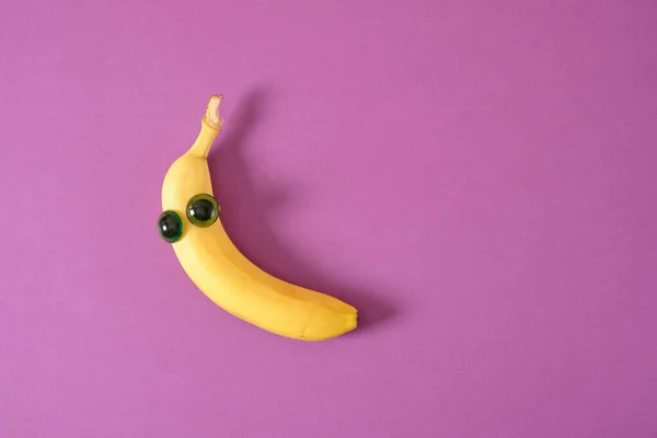 two banana face with eyes, carved cocetti merry face made of plastic doll eyes and a fresh yellow banana purple background