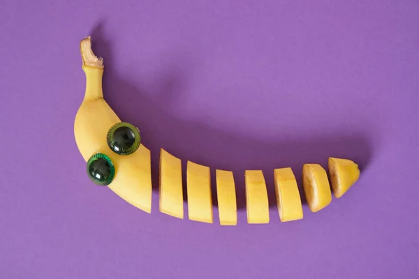 sliced banana face with eyes, carved kofetti cheerful face made of plastic doll eyes and fresh yellow banana, banana sliced directly in the peel purple background