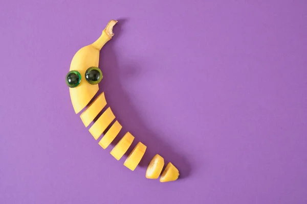 sliced banana face with eyes, carved kofetti cheerful face made of plastic doll eyes and fresh yellow banana, banana sliced directly in the peel purple background